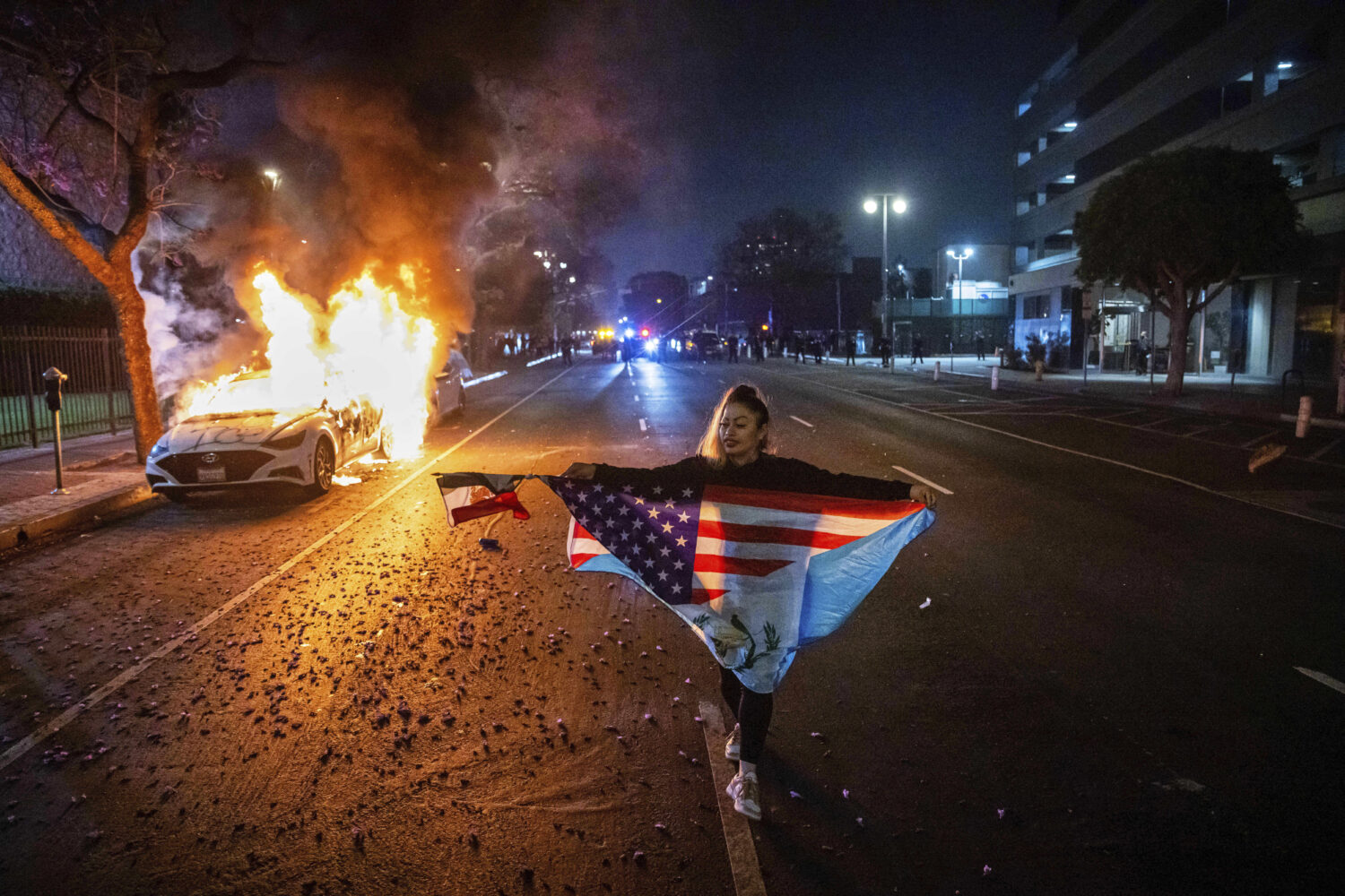 A person carrying multiple flags walks past a burning car during protests over the Trump administra...