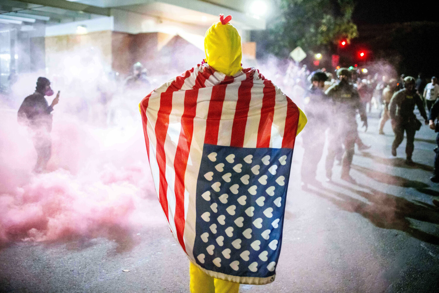 A protester, draped in an altered American flag, watches as law enforcement officers try to dispers...