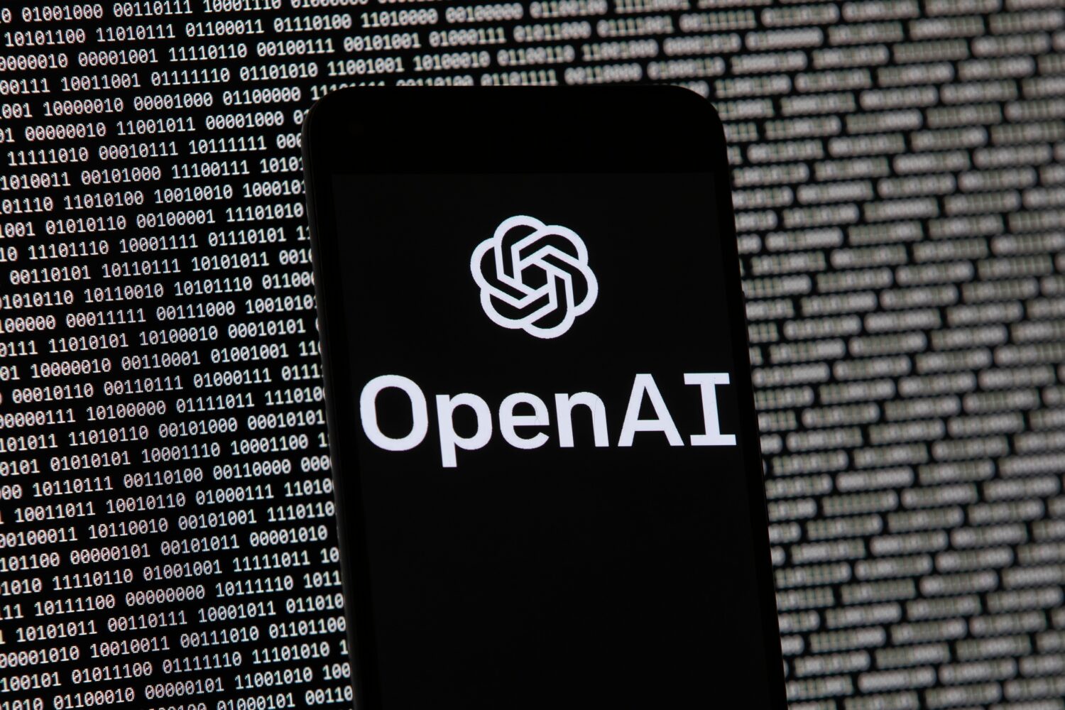 FILE - The OpenAI logo appears on a mobile phone in front of a computer screen with random binary d...