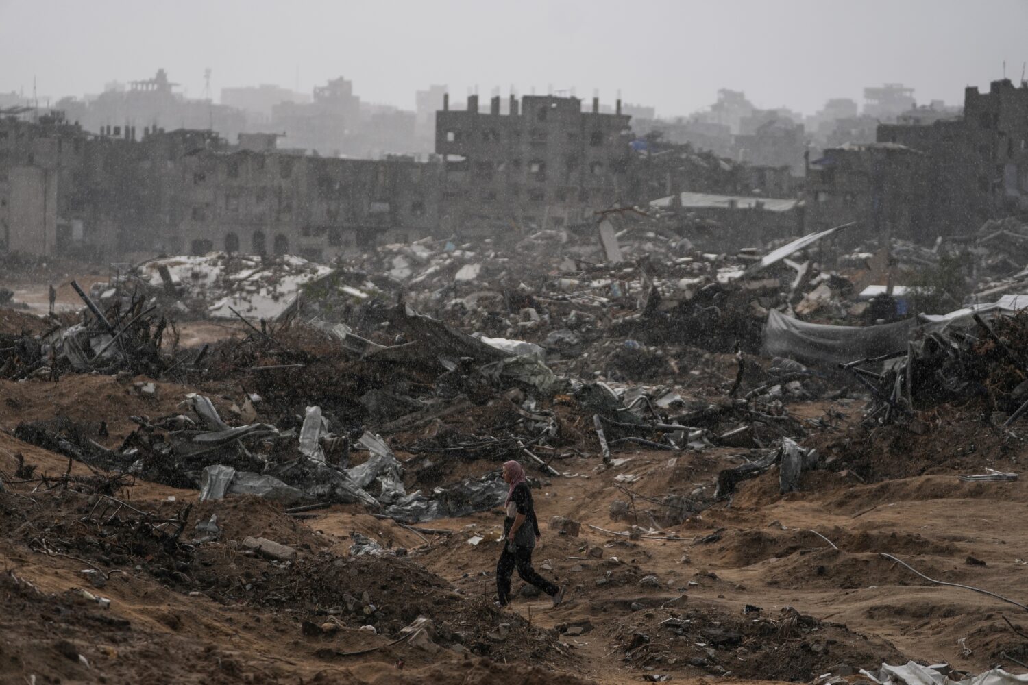 A Palestinian woman walks through a rainstorm past buildings destroyed in Israeli strikes in the Sh...