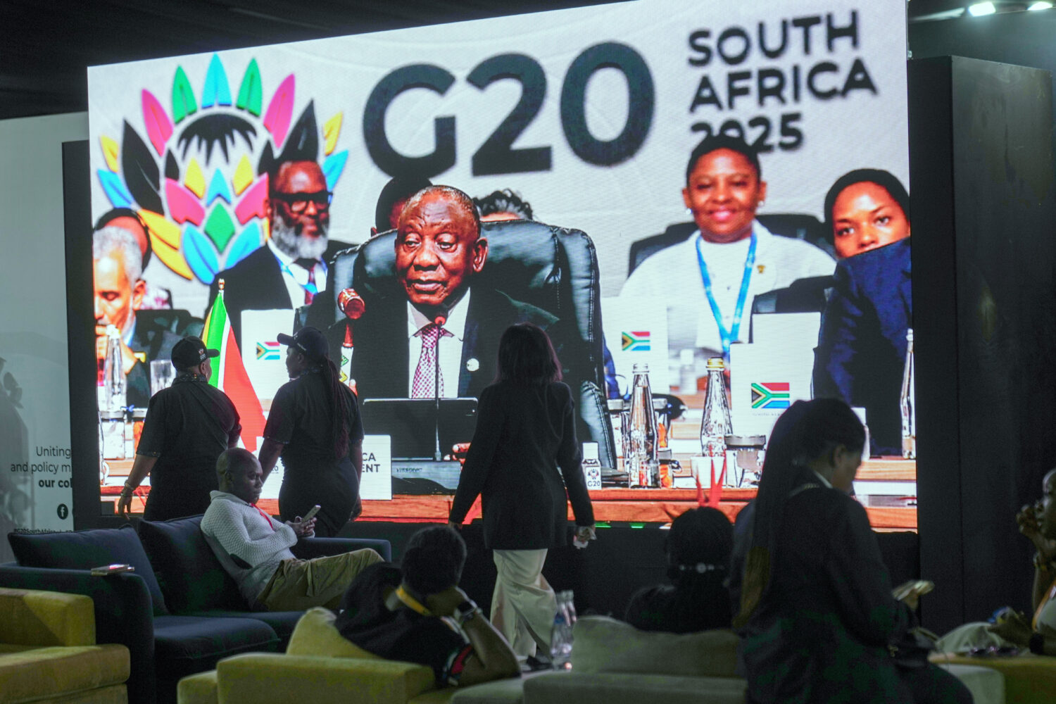 People walk by a large screen TV where South African President Cyril Ramaphosa holds a wooden gavel...