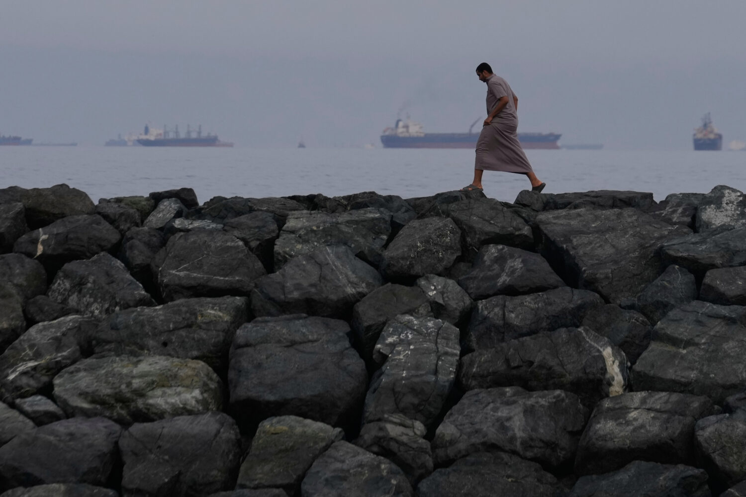 A man walks along the shore as oil tankers and cargo ships line up in the Strait of Hormuz, as seen...