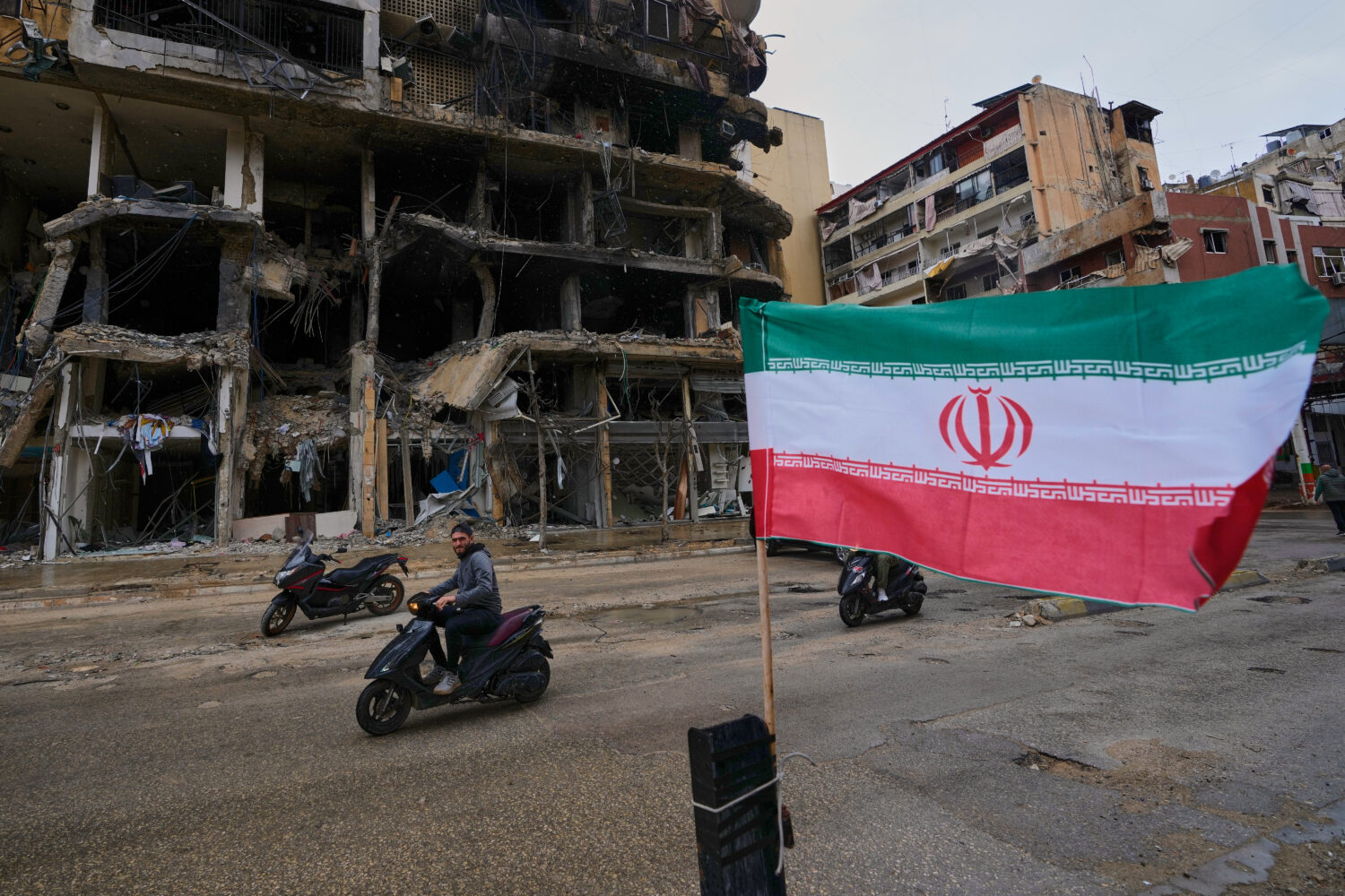 A man on his scooter passes next to an Iranian flag placed in front of a destroyed building, follow...