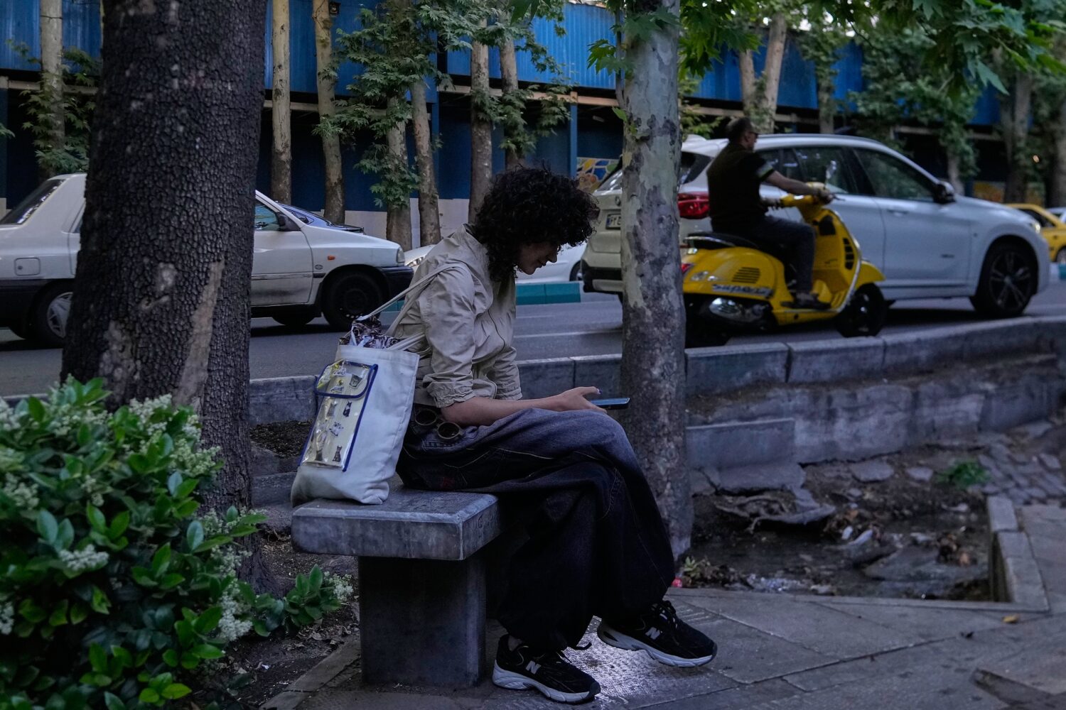 A woman checks her smartphone while sitting on a bench along a sidewalk in northern Tehran, Iran, T...