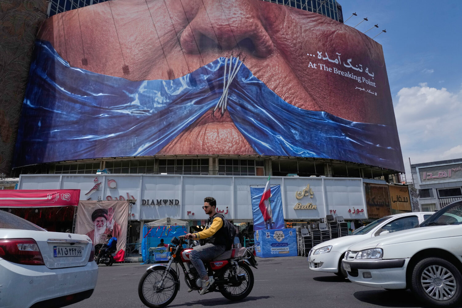 Vehicles drive past a billboard with graphic showing Strait of Hormuz and sewn lips of U.S. Preside...
