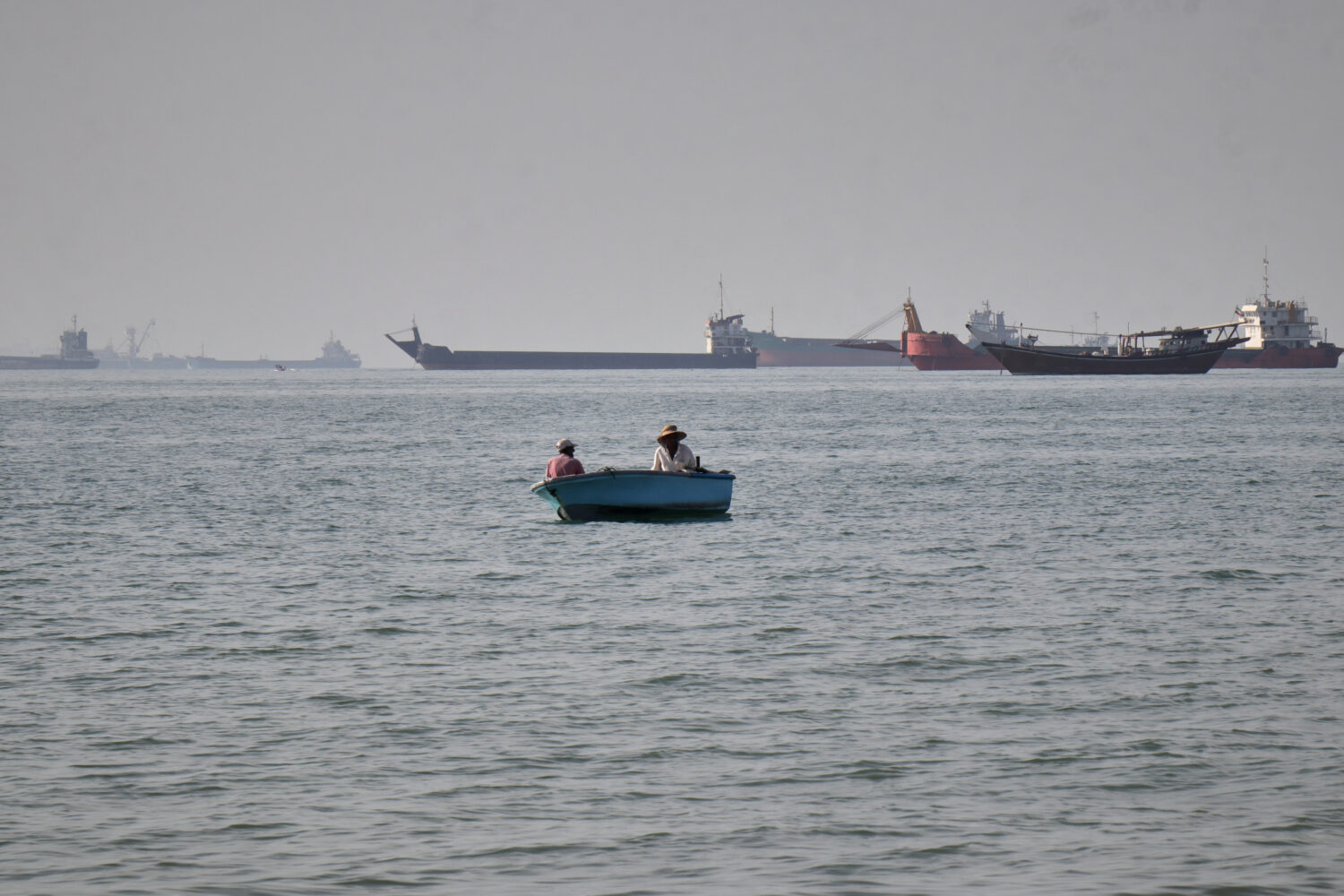 Two men sit in a small boat on the water as a mix of bulk carriers, cargo ships, and service vessel...