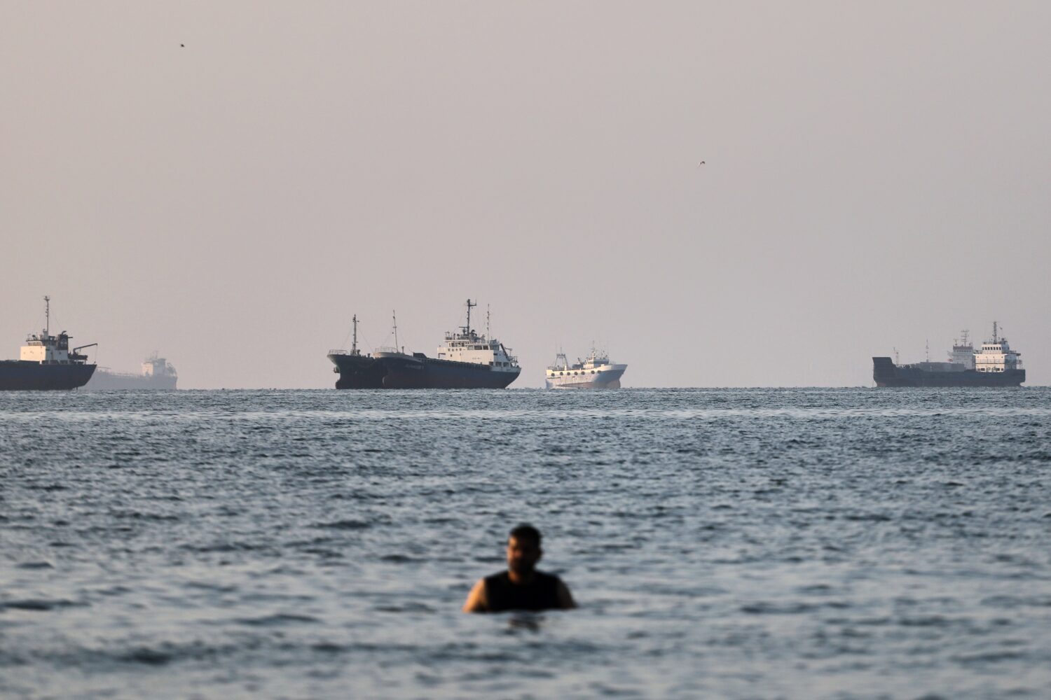 A person stands in shallow water as cargo and commercial vessels are anchored in the Strait of Horm...