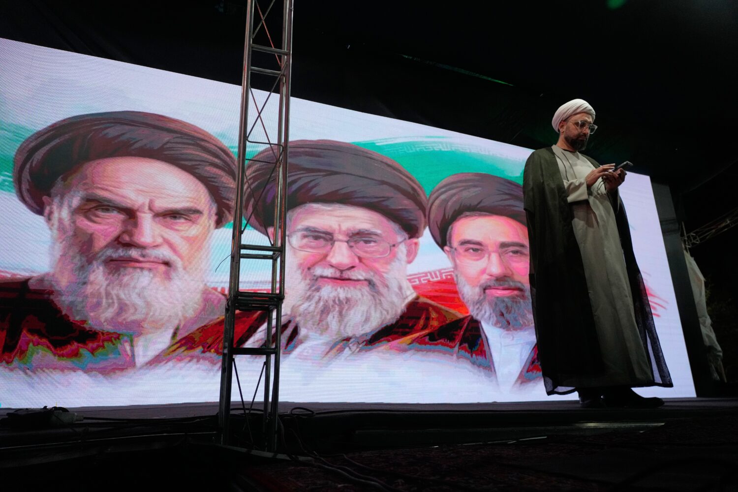 A cleric checks his cell phone on stage in front of a screen displaying portraits of the late Irani...