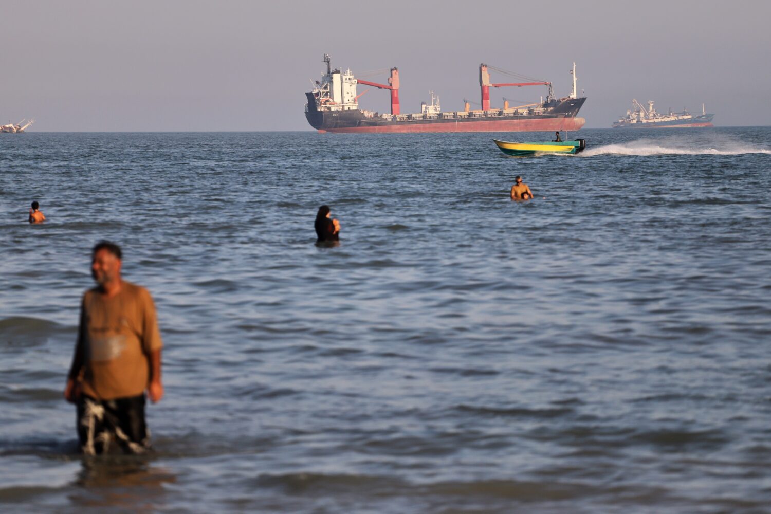 Residents swim in the waters of the Strait of Hormuz as a small motorboat passes cargo ships and ot...