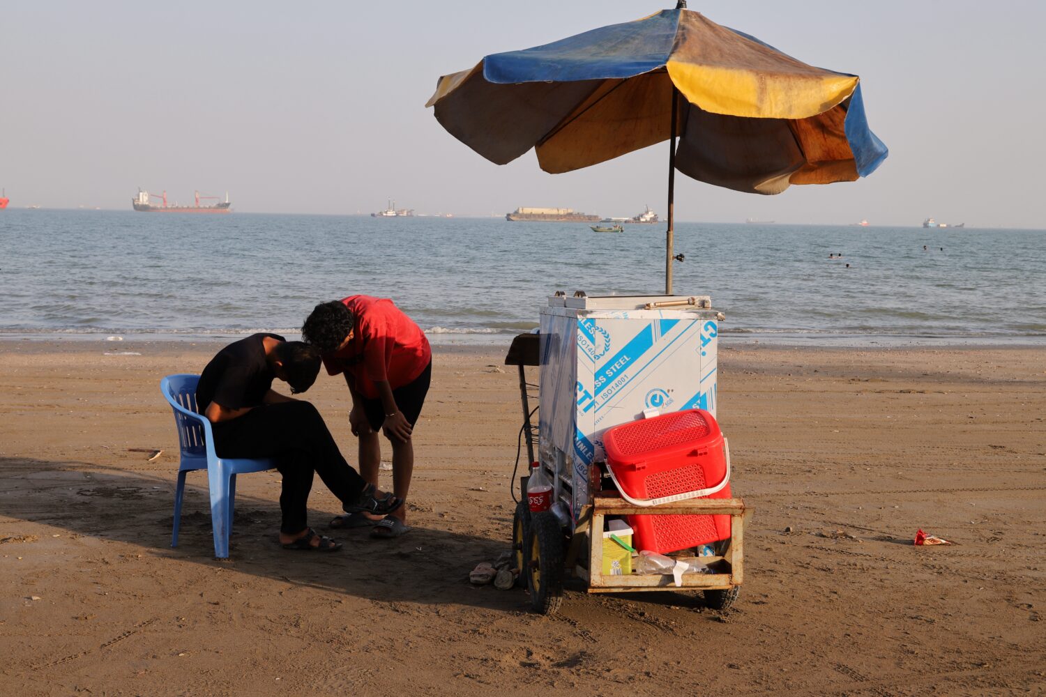 People stand next to a vendor's cart on the beach as cargo ships and offshore service vessels are a...