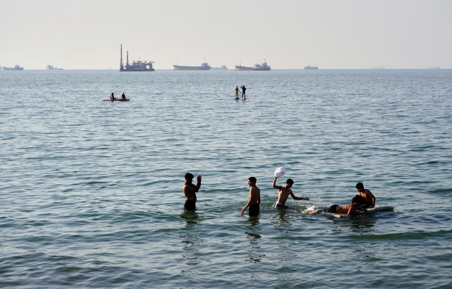 Residents swim and play in the waters of the Strait of Hormuz while cargo ships and commercial vess...