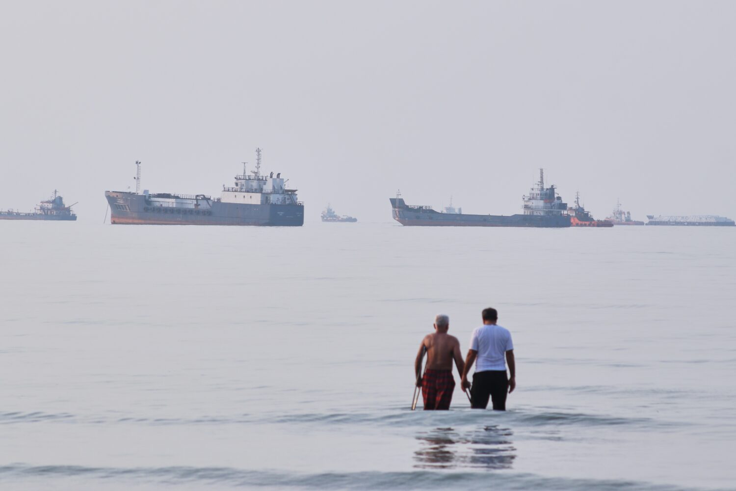 Two men wade in the waters of the Strait of Hormuz with vessels anchored in the background, off Ban...