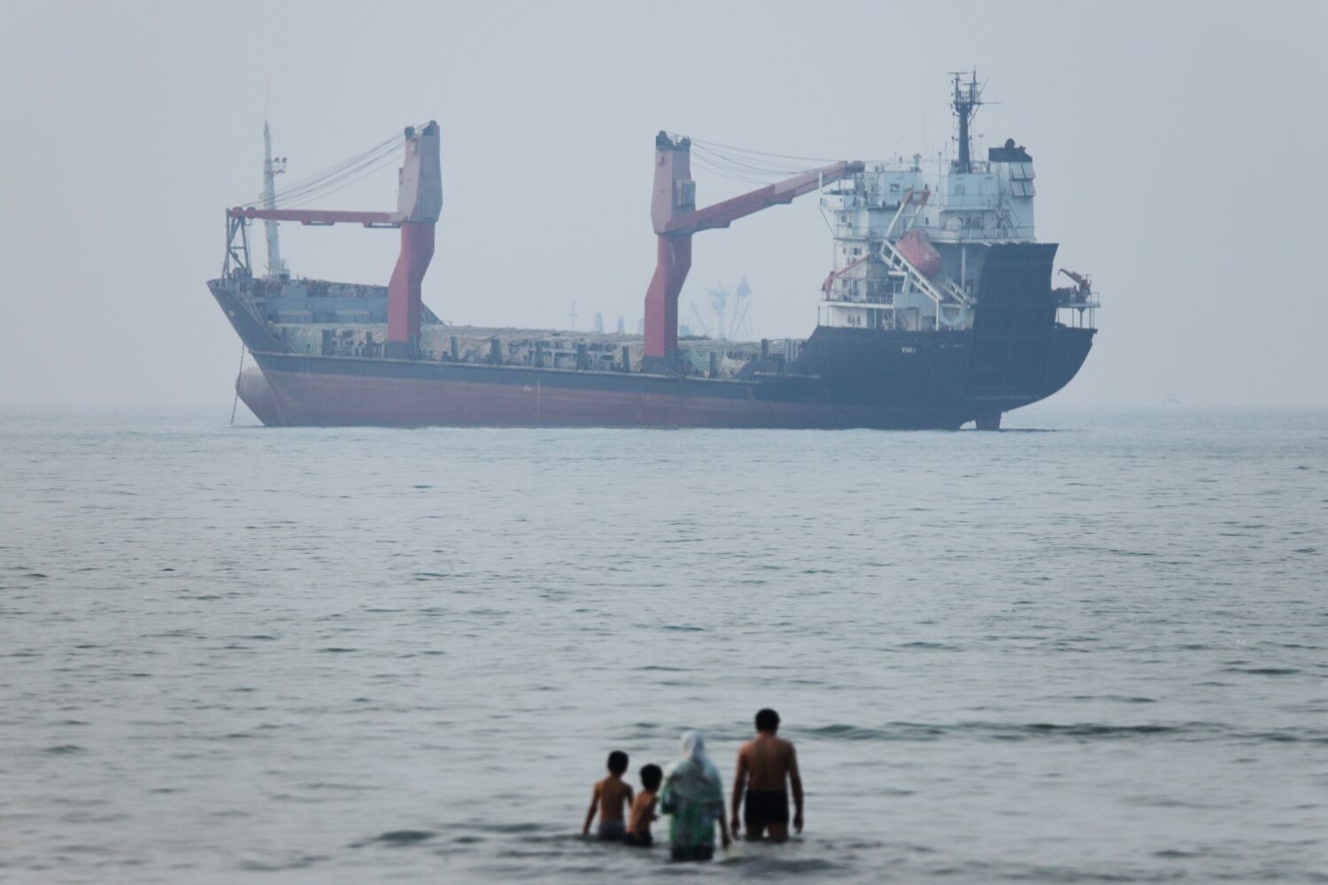 A group of people stands in shallow water as a cargo ship appears anchored in the Strait of Hormuz ...